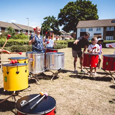 Image shows adults and children playing drums outside