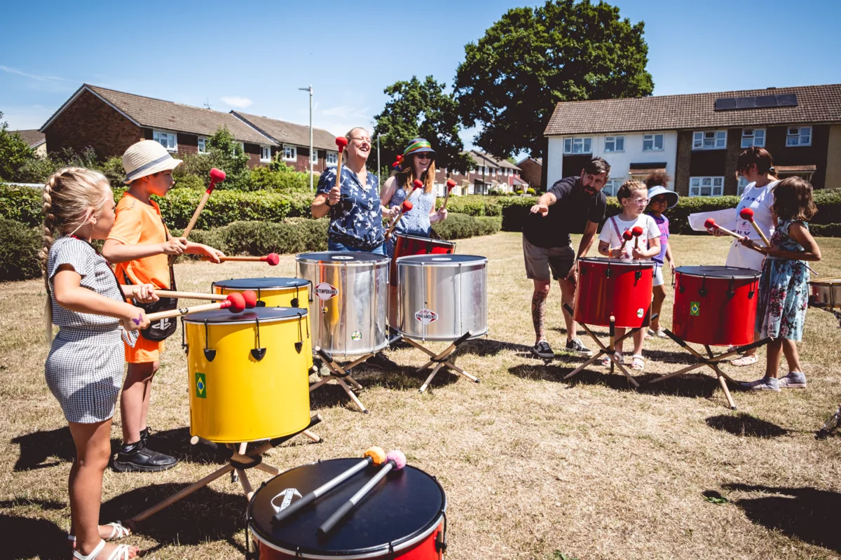 Image shows adults and children playing drums outside